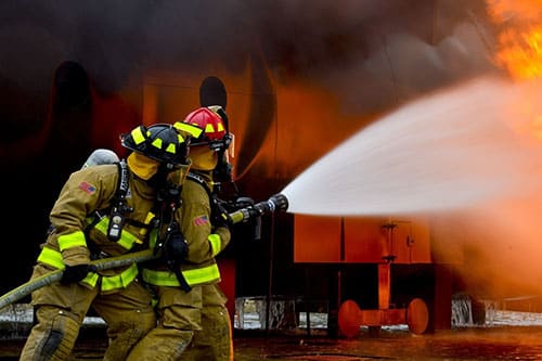 "A firefighter in high-visibility clothing stands next to a fire engine on an asphalt road. Gas flames can be seen in the background while the firefighter wears a helmet and prepares for action.
