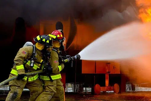 "A firefighter in high-visibility clothing stands next to a fire engine on an asphalt road. Gas flames can be seen in the background while the firefighter wears a helmet and prepares for action.